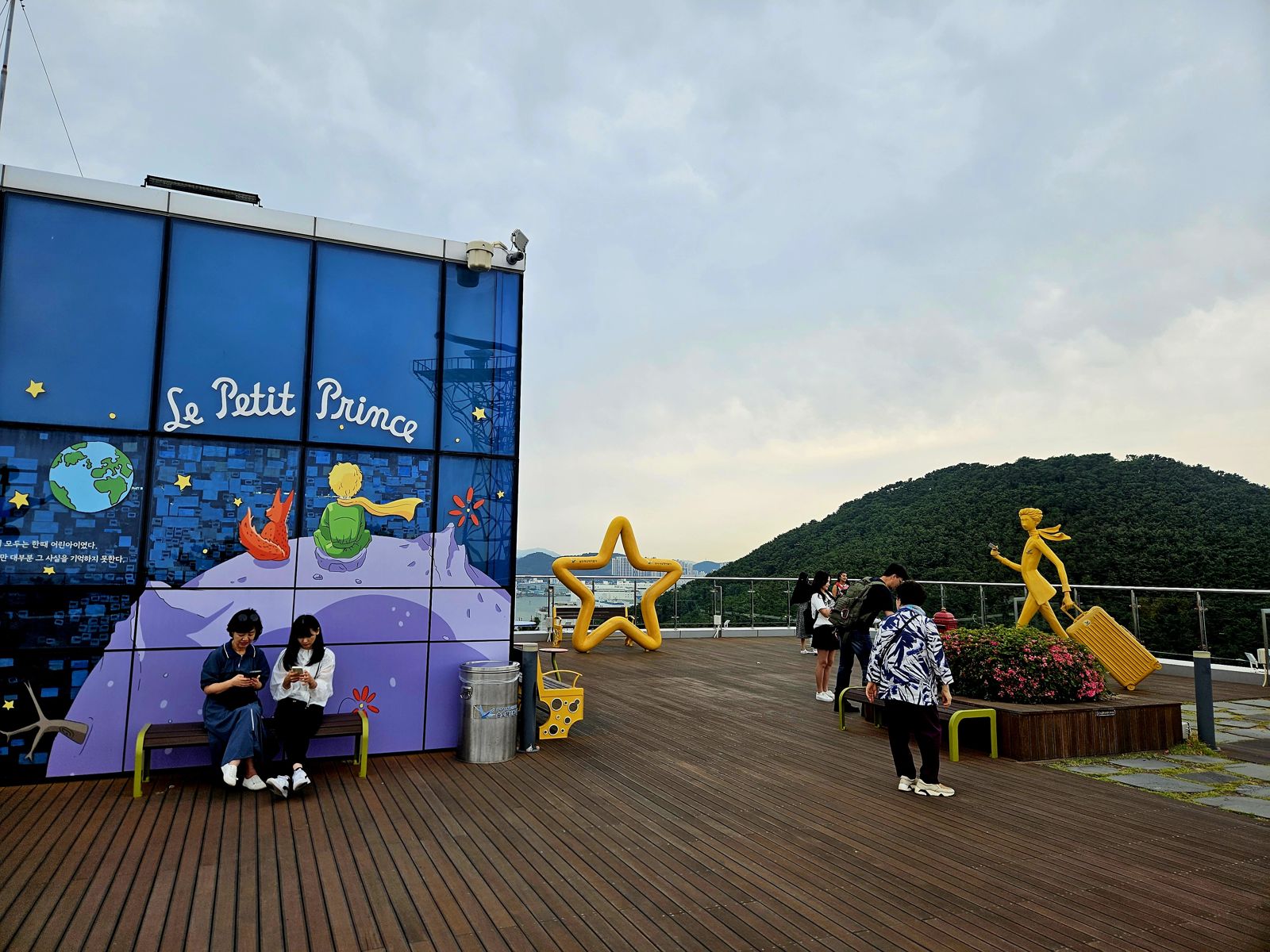Une illustration inspirée du Petit Prince sur un mur, avec deux personnages assis, entourée de visiteurs et d'une sculpture en jaune représentant une jeune fille. Le décor offre une vue sur une colline verte en arrière-plan.