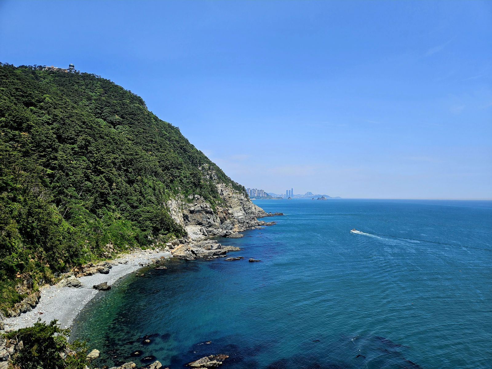 Vue panoramique d'une côte coréenne avec des collines verdoyantes, des rochers et une mer bleue. Au loin, des gratte-ciels sont visibles sous un ciel clair.