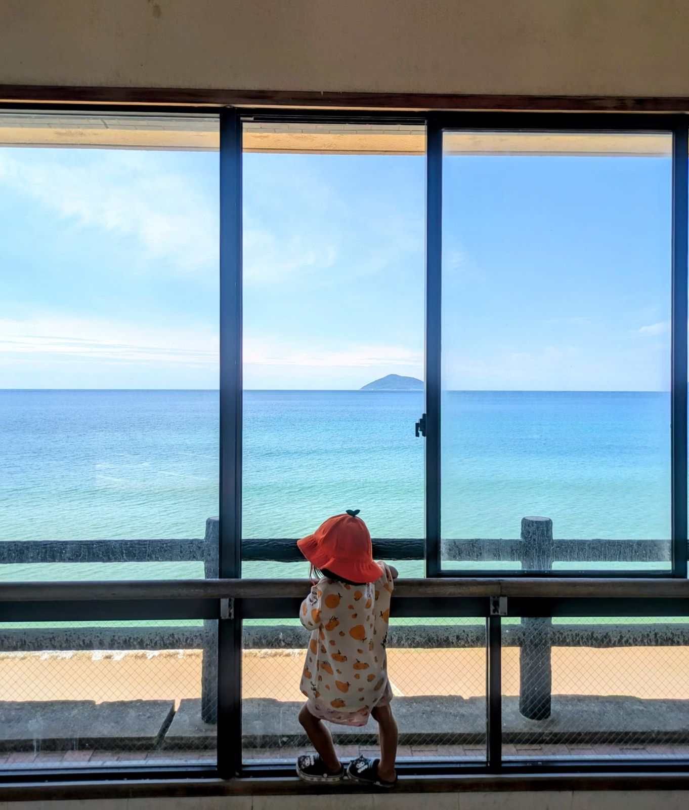 Un enfant avec un chapeau orange se tient debout devant une grande fenêtre, regardant la mer paisible et une petite île au loin.