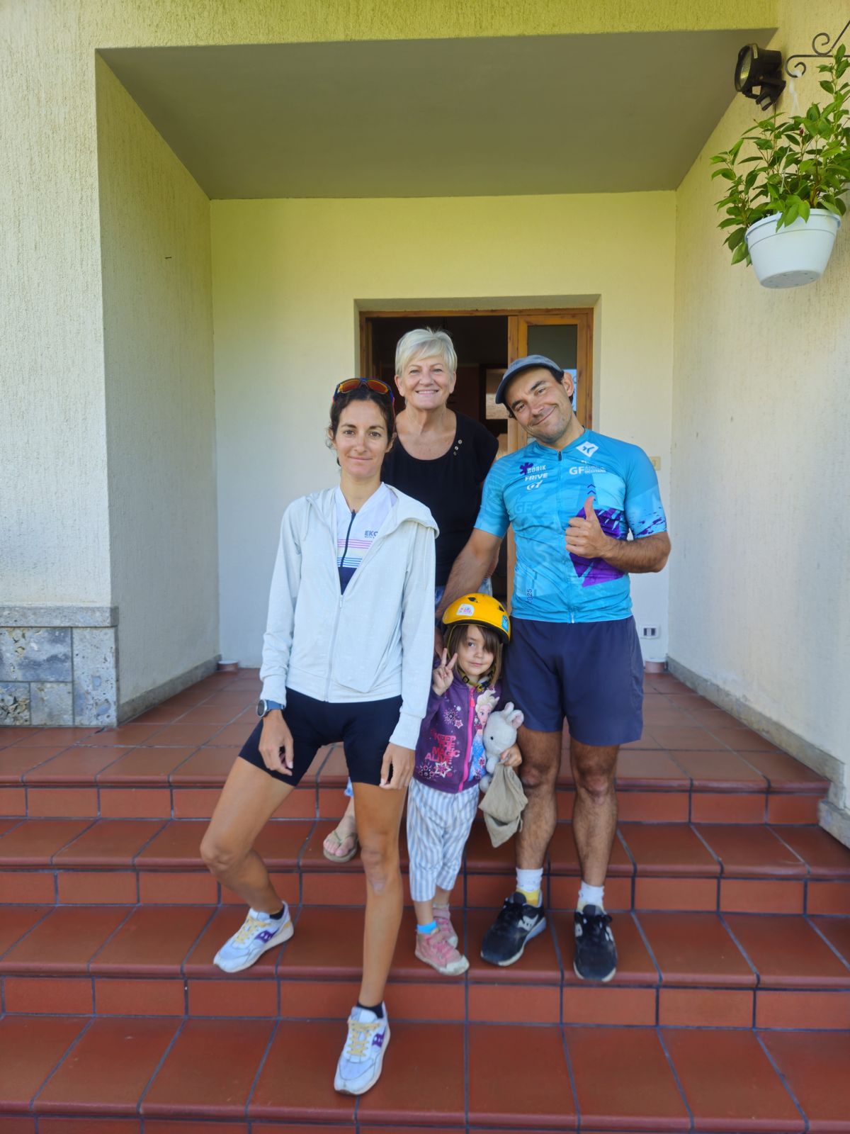 Quatre personnes posant devant une maison, souriant à la caméra. Un homme et une femme adultes debout, avec une petite fille au centre portant un casque de vélo. Une autre femme est derrière eux.