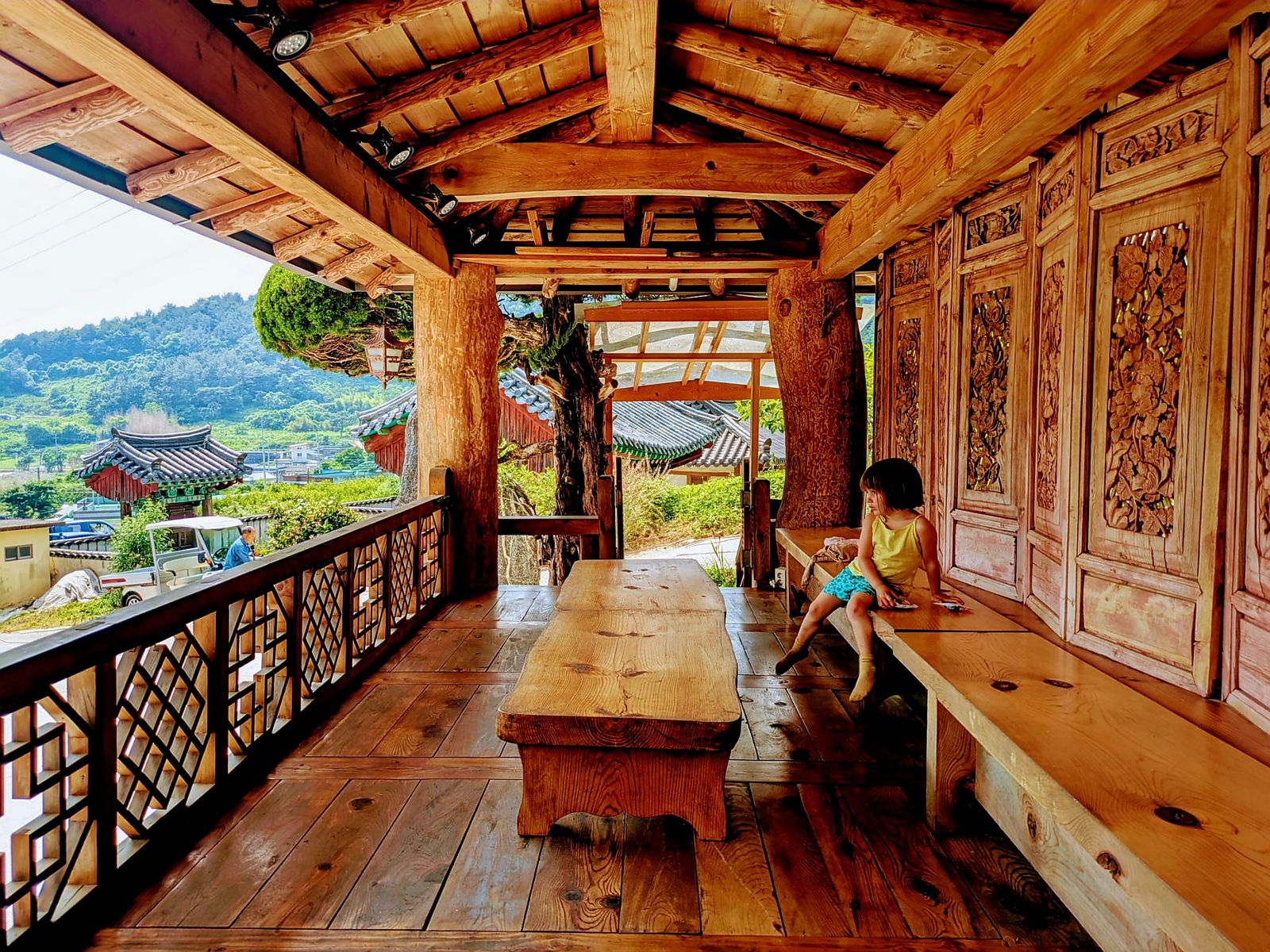 Une petite fille assise sur un banc en bois dans un café traditionnel coréen, entourée de murs en bois sculpté et avec des montagnes verdoyantes en arrière-plan.