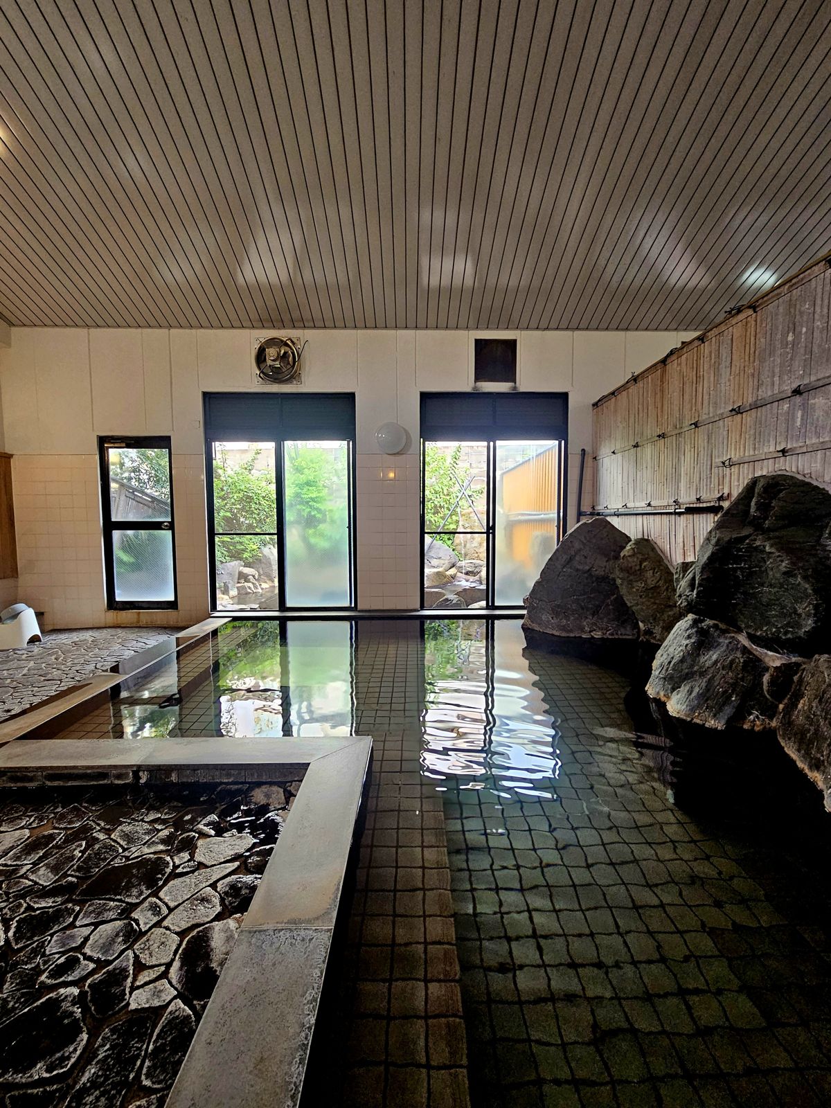 Intérieur d'un onsen avec des bassins d'eau chaude, entouré de rochers et de grandes fenêtres donnant sur un jardin.