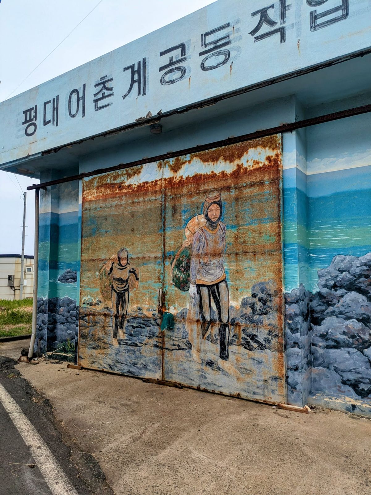 Mur peint représentant des pêcheurs sur une porte en métal rouillé, avec le ciel et la mer en arrière-plan.