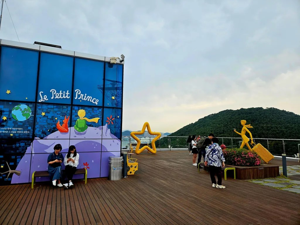 Une illustration inspirée du Petit Prince sur un mur, avec deux personnages assis, entourée de visiteurs et d'une sculpture en jaune représentant une jeune fille. Le décor offre une vue sur une colline verte en arrière-plan.