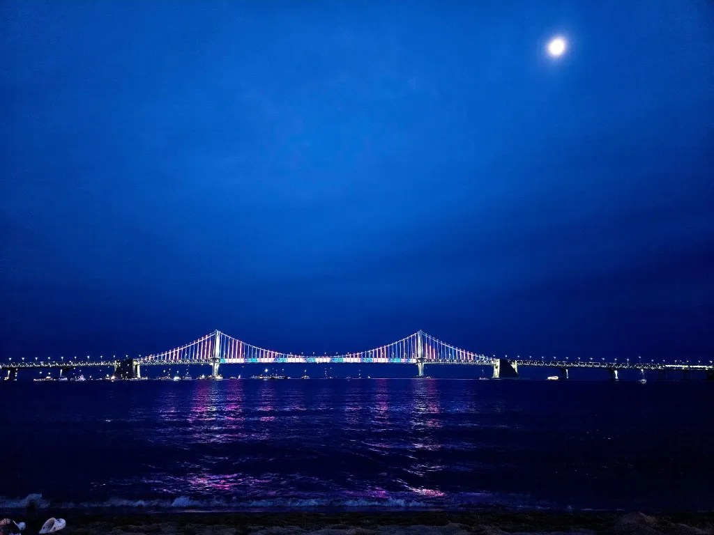 Vue nocturne du pont Gwangan à Busan illuminé par des lumières colorées, avec la lune visible dans le ciel.
