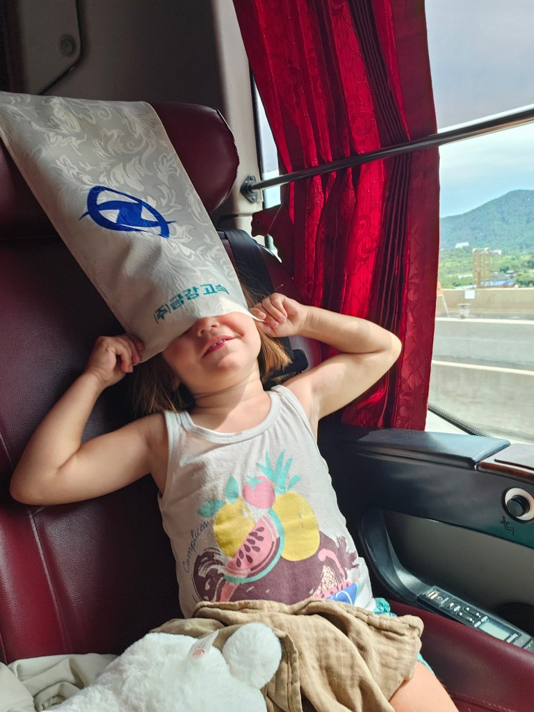 Une jeune fille assise dans un bus, portant un bonnet sur la tête, avec un air joyeux et étonné, dans un environnement de sièges rouges et rideaux.