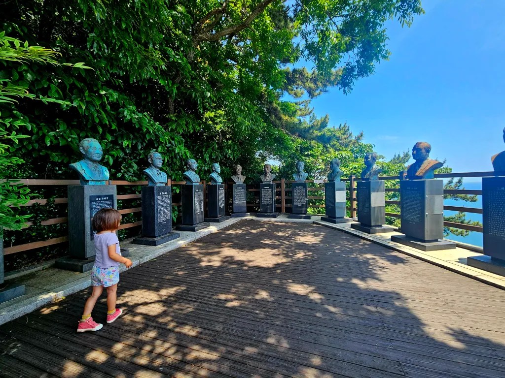 Une petite fille marche sur une plateforme en bois, entourée de bustes monumentaux de personnalités. L'environnement est verdoyant avec des arbres en arrière-plan et un ciel bleu.