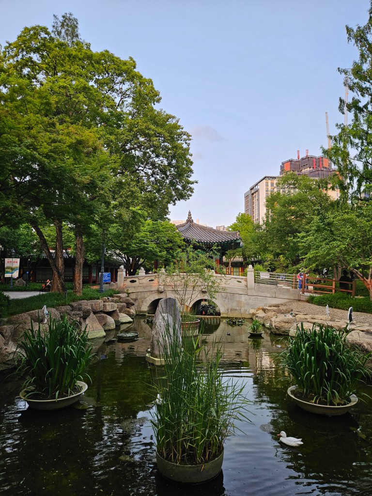 Un parc avec un petit étang, des plantes aquatiques et un joli pont en pierre, entouré d'arbres luxuriants et d'un bâtiment traditionnel en arrière-plan.