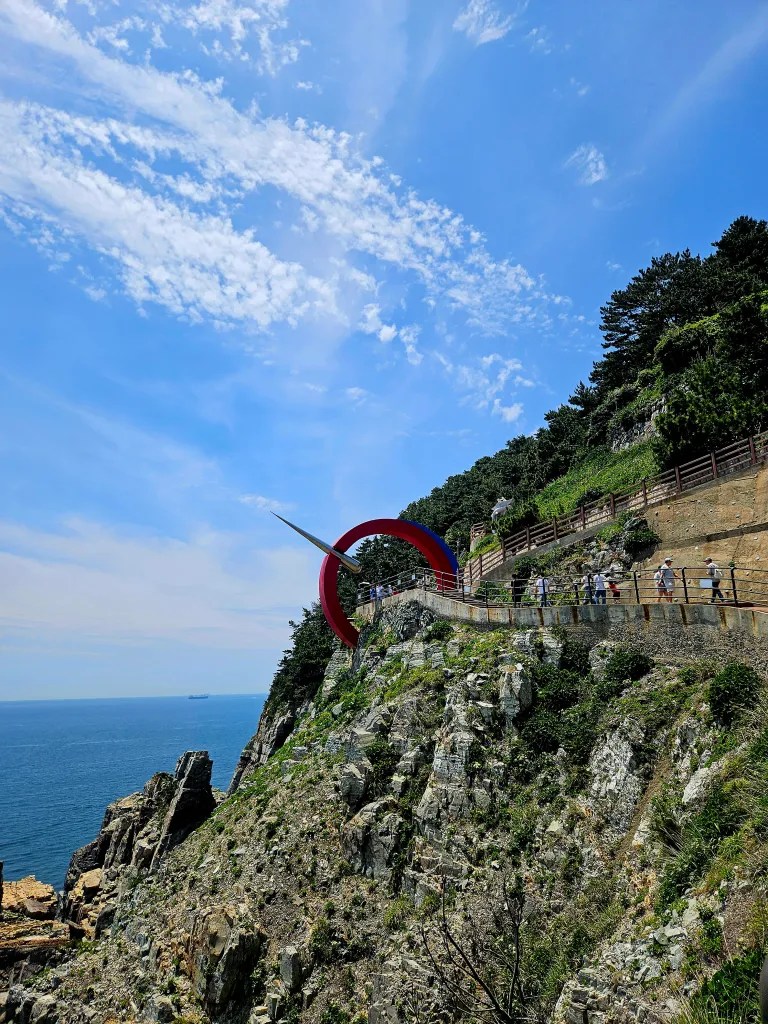 Un belvédère avec une sculpture rouge surplombant la mer, entouré d'une falaise rocheuse et d'une végétation verdoyante, sous un ciel bleu parsemé de nuages.