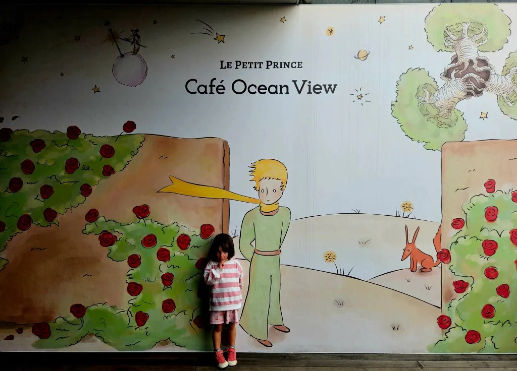 Une petite fille se tient devant une murale colorée représentant Le Petit Prince, avec des roses et un renard, dans un café nommé 'Café Ocean View'.