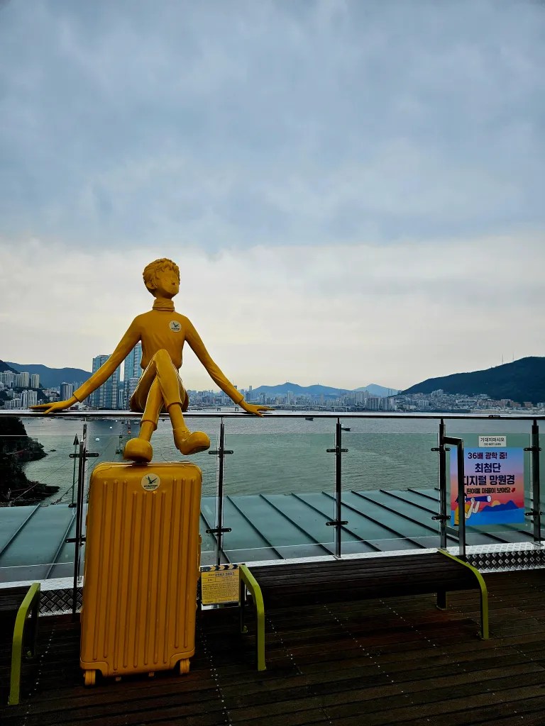 Statue jaune d'un enfant assis sur une valise, avec vue sur la mer et la ville en arrière-plan.