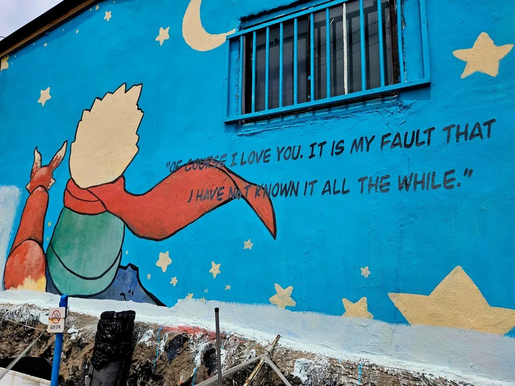 Mur peint représentant le Petit Prince tenant une rose, avec une citation écrite en anglais sur fond bleu, parsemé d'étoiles.
