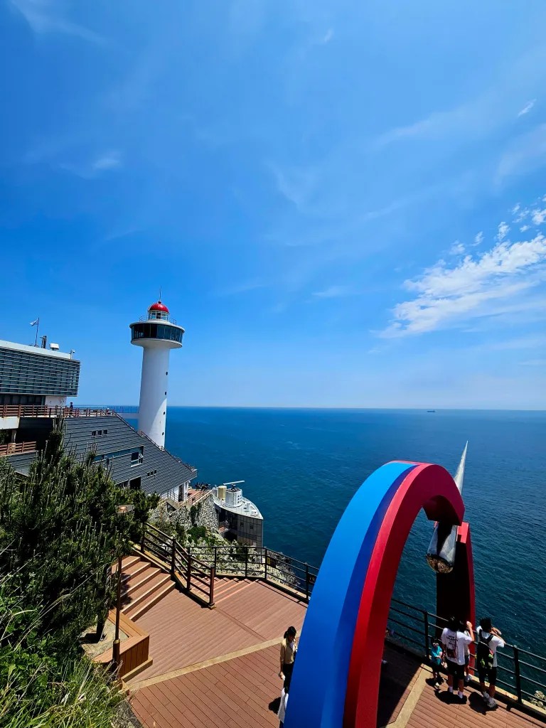 Vue panoramique d'un phare avec un ciel bleu clair et l'océan en arrière-plan, des touristes admirant le paysage près d'un arc coloré.