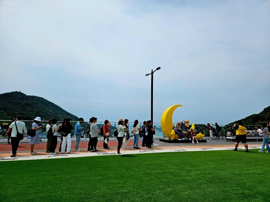 Un groupe de personnes fait la queue pour prendre des photos avec des statues inspirées du Petit Prince, au bord de la mer, avec des collines en arrière-plan.