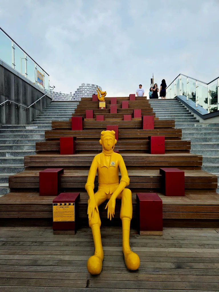 Une sculpture en jaune représentant un personnage assis sur des marches en bois, entourée de blocs rouges, avec des visiteurs en arrière-plan.