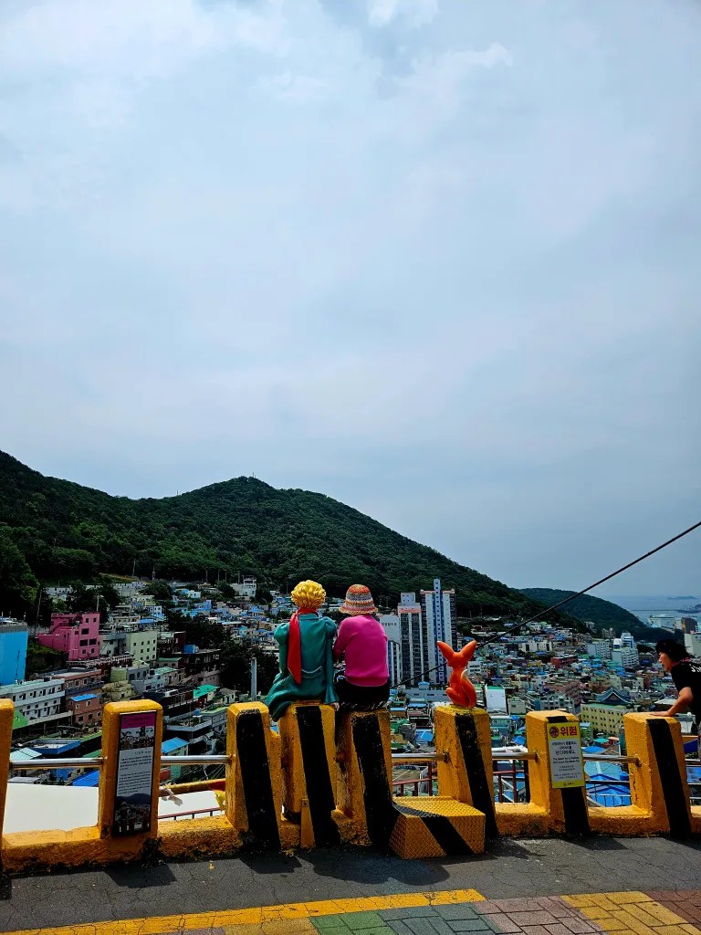Vue panoramique de Busan avec des personnages de statues inspirés du Petit Prince, surplombant des maisons colorées et des collines verdoyantes.