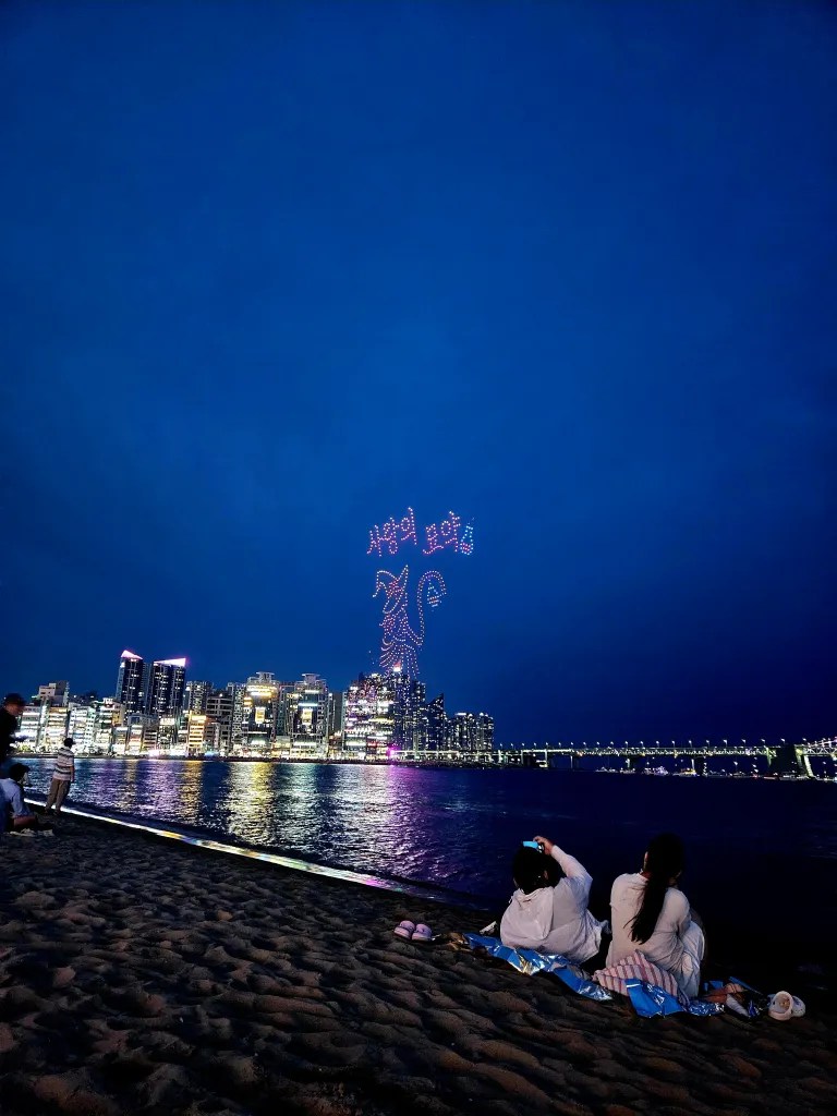 Une vue de la plage de Busan la nuit, avec des spectateurs assis sur le sable, observant un spectacle de drones qui illuminent le ciel avec des formes colorées et des lumières vives, tandis que la ville se dresse en arrière-plan.