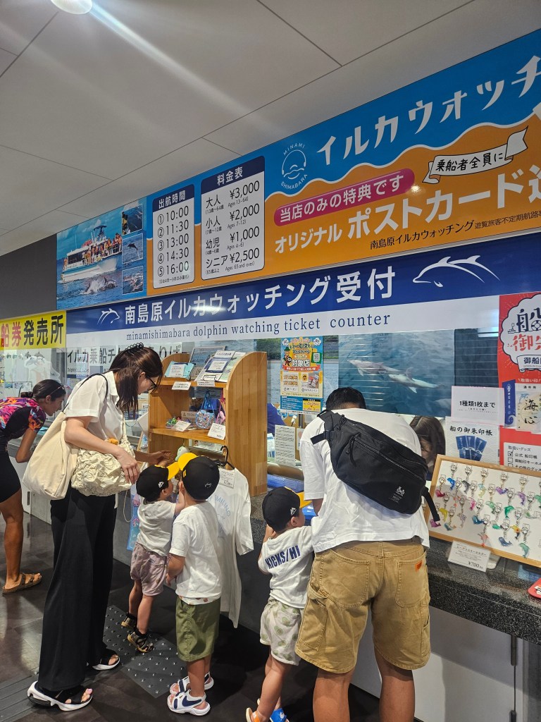 Une famille se tient devant un comptoir de vente de billets pour des excursions d'observation de dauphins. On peut voir des panneaux d'affichage avec des horaires et des prix, ainsi que des brochures et des souvenirs. Les enfants, portant des casquettes, semblent intéressés par les informations affichées.