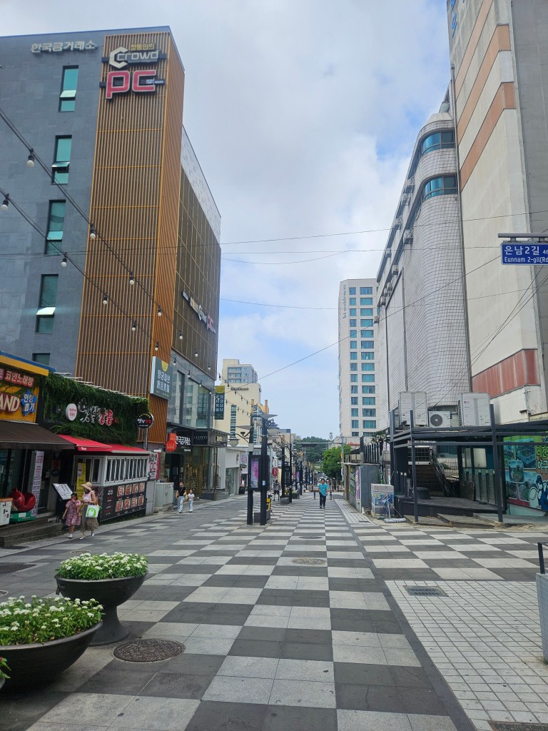Vue d'une rue moderne et peu fréquentée à Jeju, avec des bâtiments commerciaux et des espaces verts, sous un ciel partiellement nuageux.