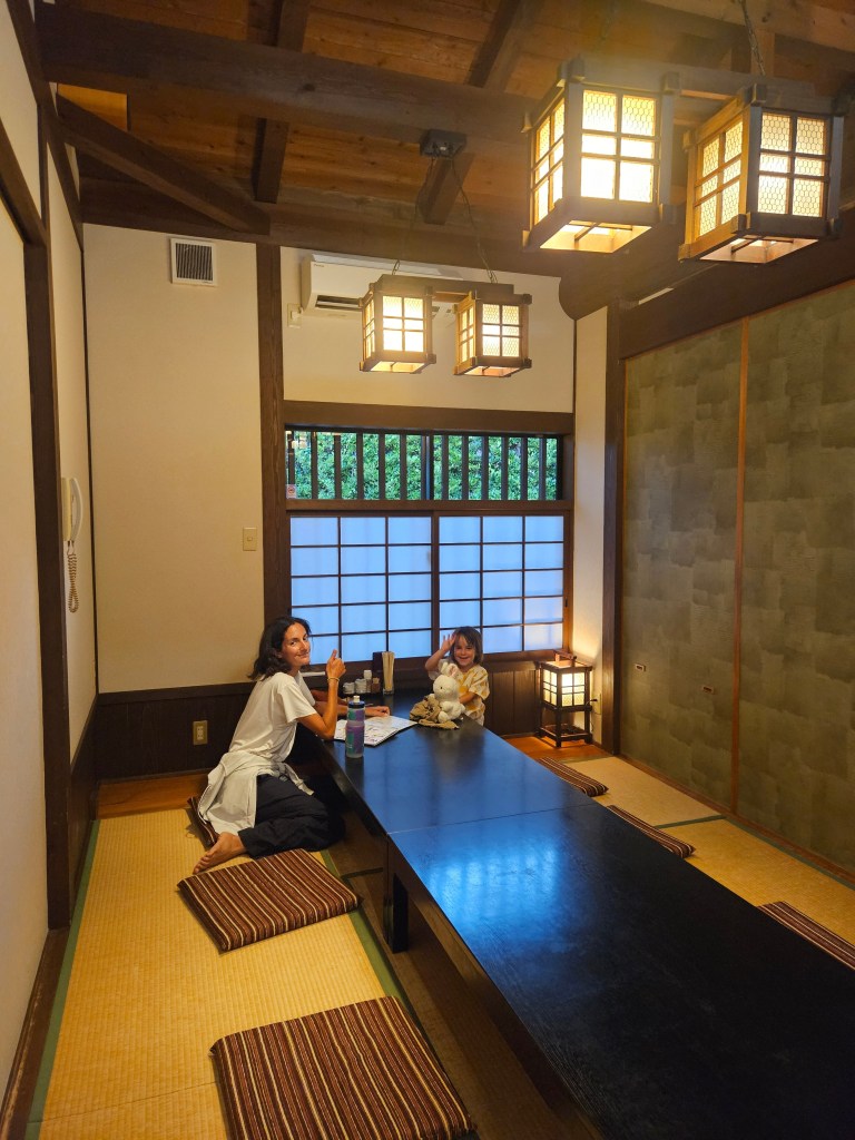 Intérieur d'une auberge japonaise traditionnelle avec un sol en tatami, où une femme et une enfant sont assises à une grande table en bois sombre, entourées de lanternes décoratives.