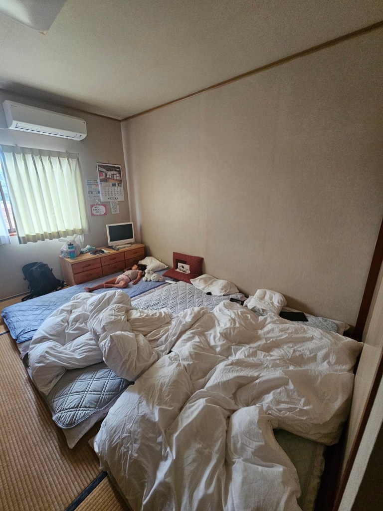 Chambre d'hôtel japonaise avec deux matelas et des couvertures en désordre, une petite télévision et une fenêtre laissant entrer la lumière.