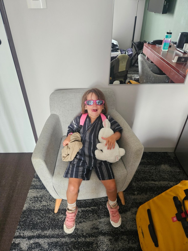 Une petite fille assise sur un fauteuil dans une chambre d'hôtel, portant des lunettes de soleil, un vêtement en tissu léger, et tenant un doudou et un vêtement plié.