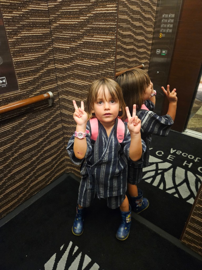 Une petite fille portant un yukata rayé et des bottes, se tenant dans un ascenseur tout en montrant deux doigts en signe de paix. Elle porte un petit sac à dos rose et a l'air joyeuse.