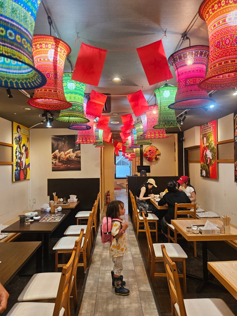 Intérieur coloré d'un restaurant avec des lanternes décoratives et des drapeaux. Une petite fille avec un sac à dos se tient debout au centre, regardant autour d'elle.