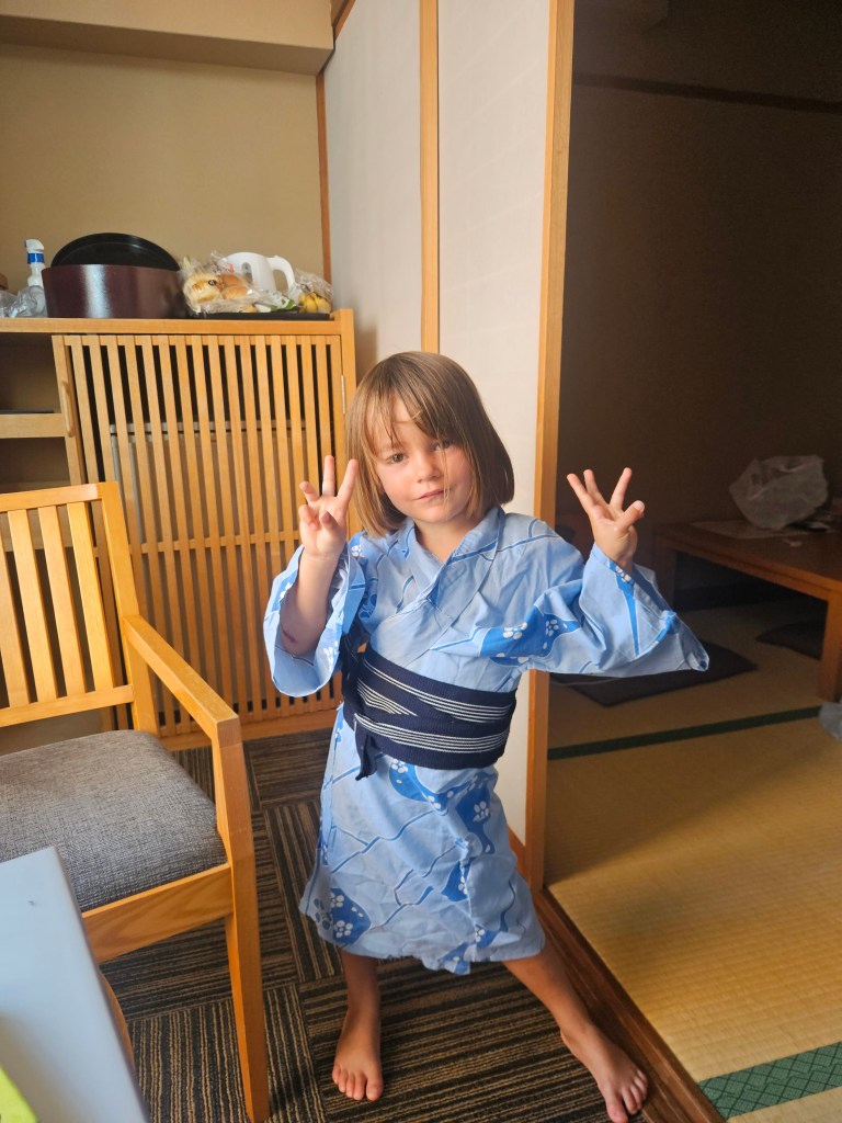 Une petite fille en kimono bleu avec des motifs japonais, se tenant dans une chambre d'hôtel, montrant des signes de paix avec ses mains.