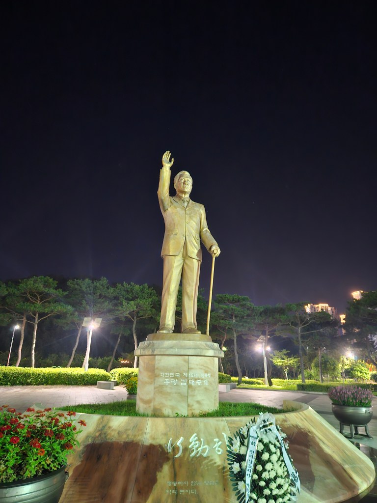 Statue d'une figure historique levant la main, éclairée par des lumières de parc la nuit, entourée de verdure et de fleurs.
