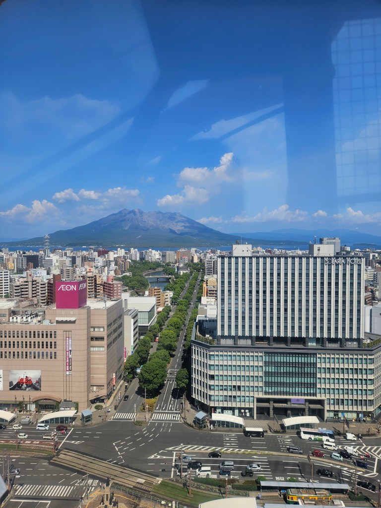 Vue panoramique de la ville de Kagoshima avec le volcan Sakurajima à l'arrière-plan et des bâtiments modernes en premier plan.