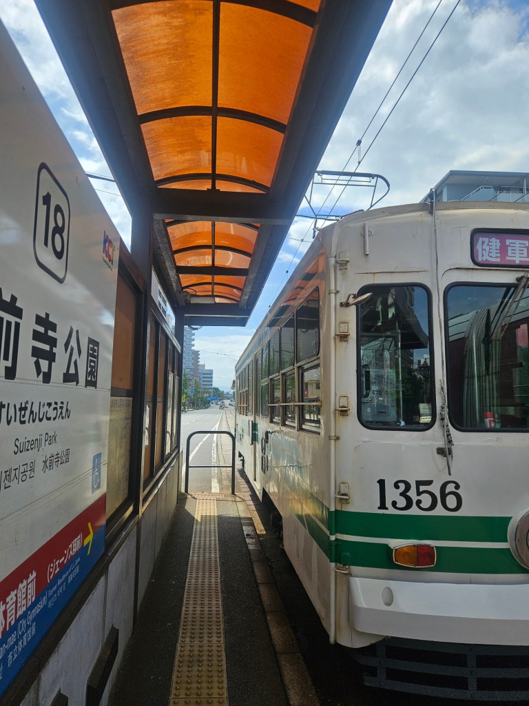 Un tramway à l'arrêt à la station Suizenji Park, avec un toit orange, sous un ciel partiellement nuageux.