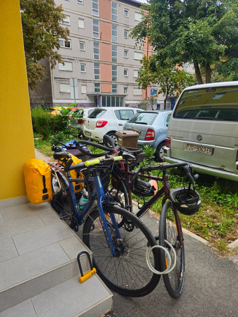 Des vélos avec des sacs de voyage, garés près d'un bâtiment résidentiel coloré, entourés de voitures et de verdure.