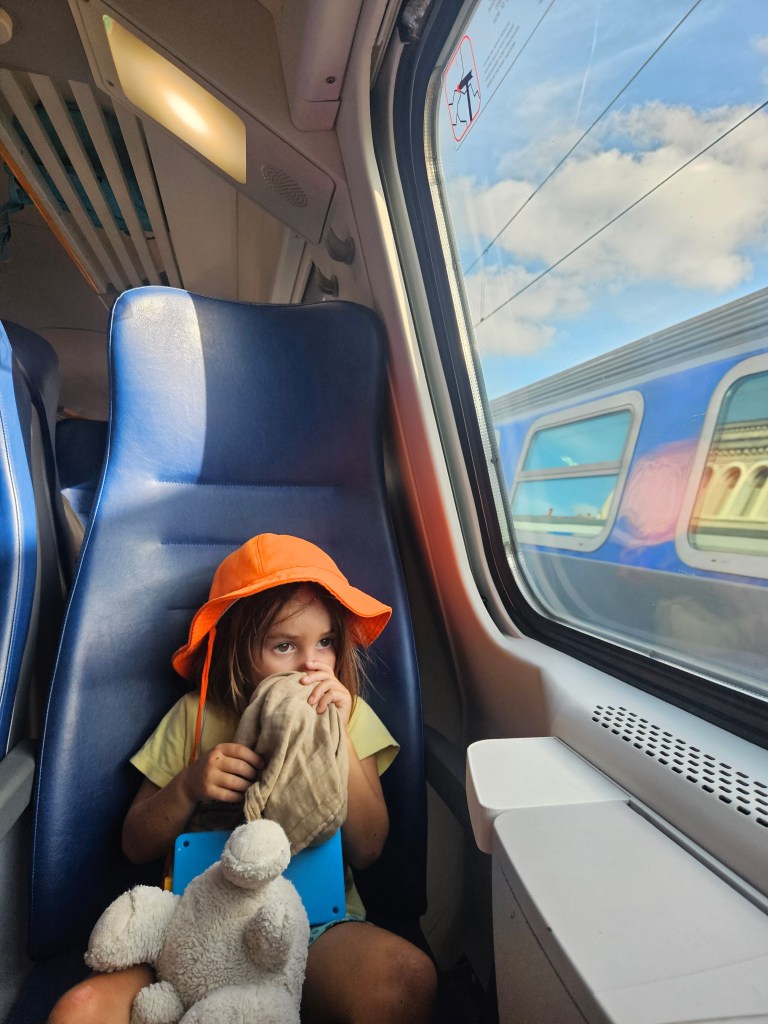 Une jeune fille portant un chapeau orange assise dans un train, tenant un doudou et regardant par la fenêtre.