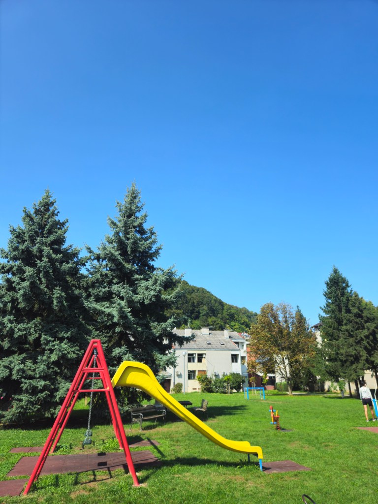 Une aire de jeux avec un toboggan jaune et une structure rouge, entourée d'arbres et d'un ciel bleu.
