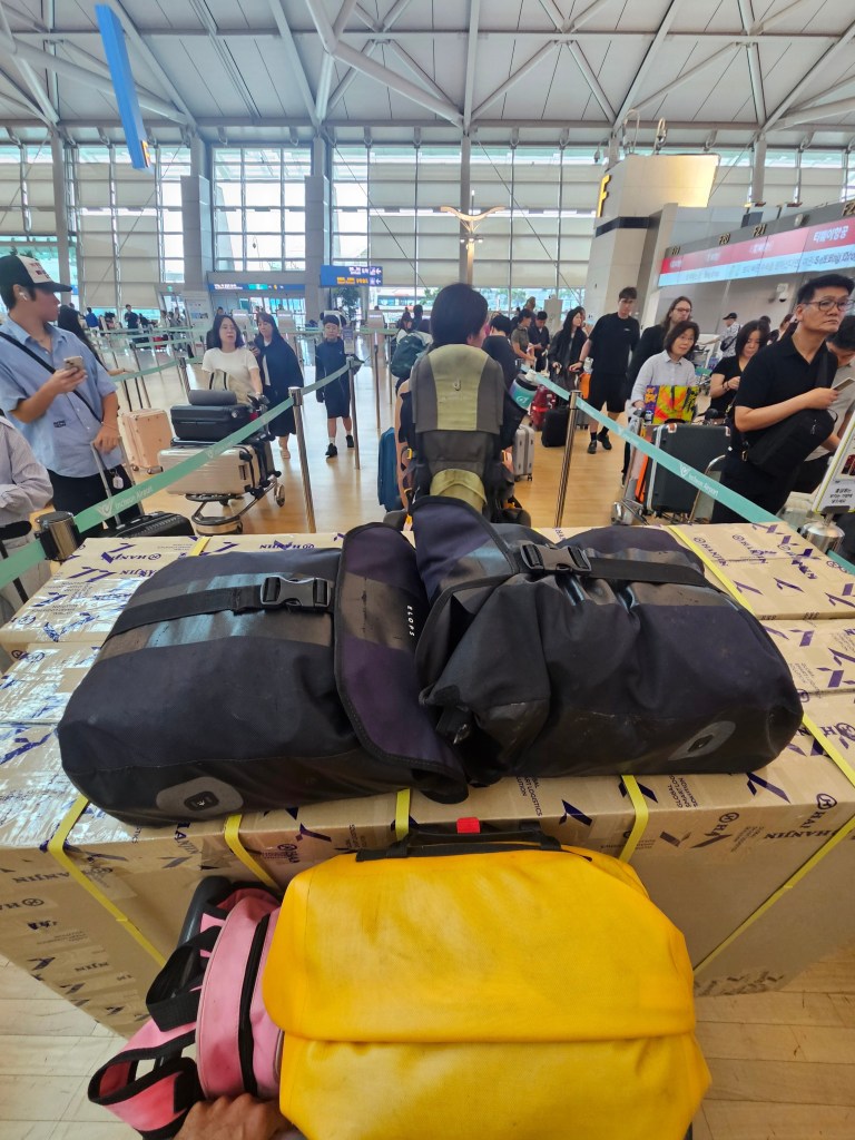 Photo d'un comptoir d'enregistrement à l'aéroport, avec des bagages empilés, dont des sacoches de vélo et un carton, dans une file d'attente.