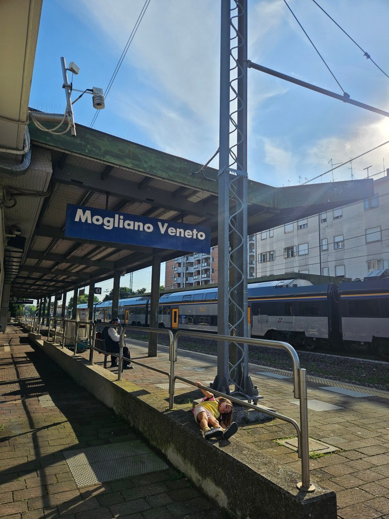 Gare de Mogliano Veneto avec le panneau indiquant le nom de la gare, des trains en arrière-plan et des personnes assises sur le banc.