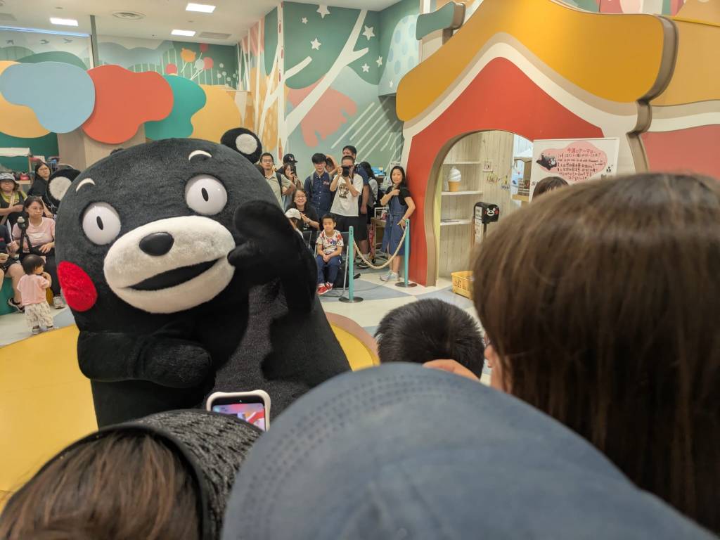 Kumamon, la mascotte de Kumamoto, interagissant avec des enfants et des visiteurs dans un espace public animé, entouré d'un public enthousiaste.