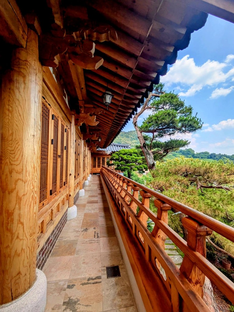 Vue d'un couloir en bois traditionnel, avec des colonnes en bois et un garde-corps, bordé d'arbres et de verdure sous un ciel bleu.