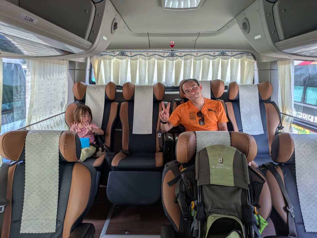 Intérieur d'un bus avec des sièges en cuir marron et noir, où un homme sourit et fait un signe de paix tandis qu'une petite fille assise à côté lui fait coucou.