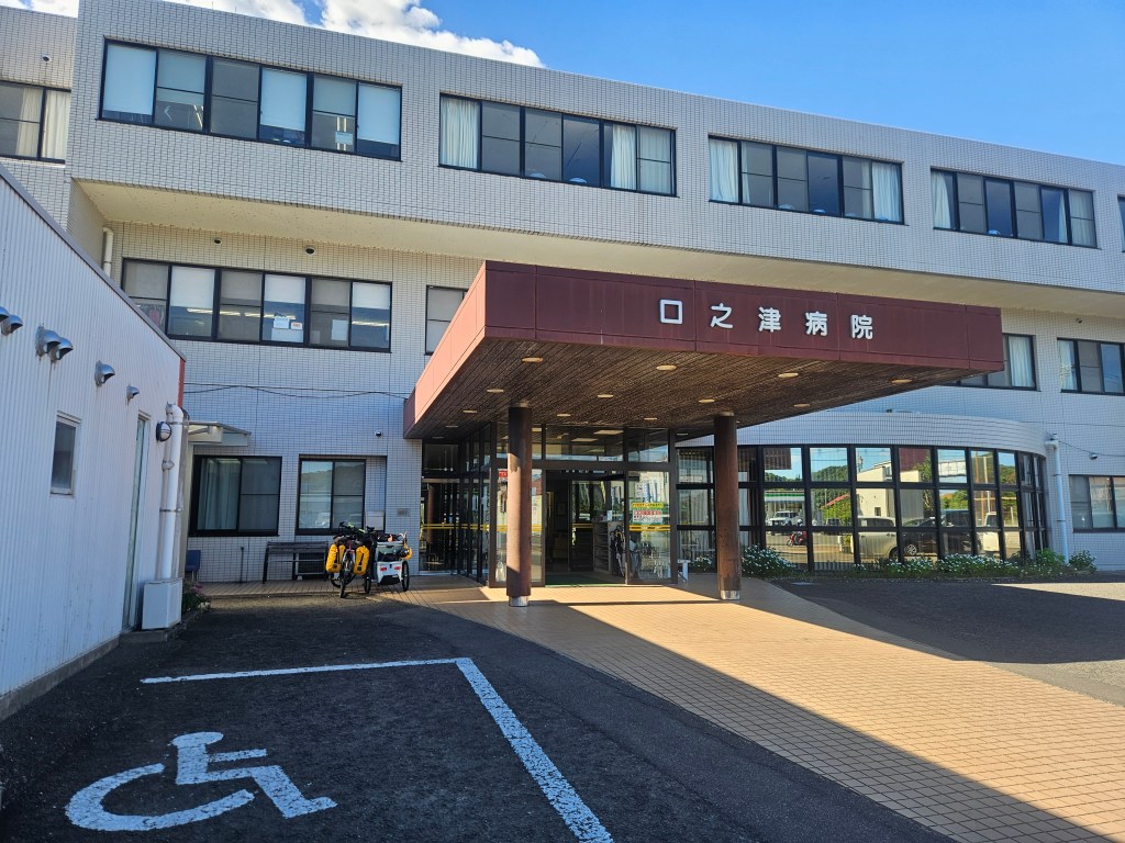 Entrée d'un hôpital avec des fleurs blanches à l'avant et des fauteuils à côté, devant une grande façade avec des fenêtres.