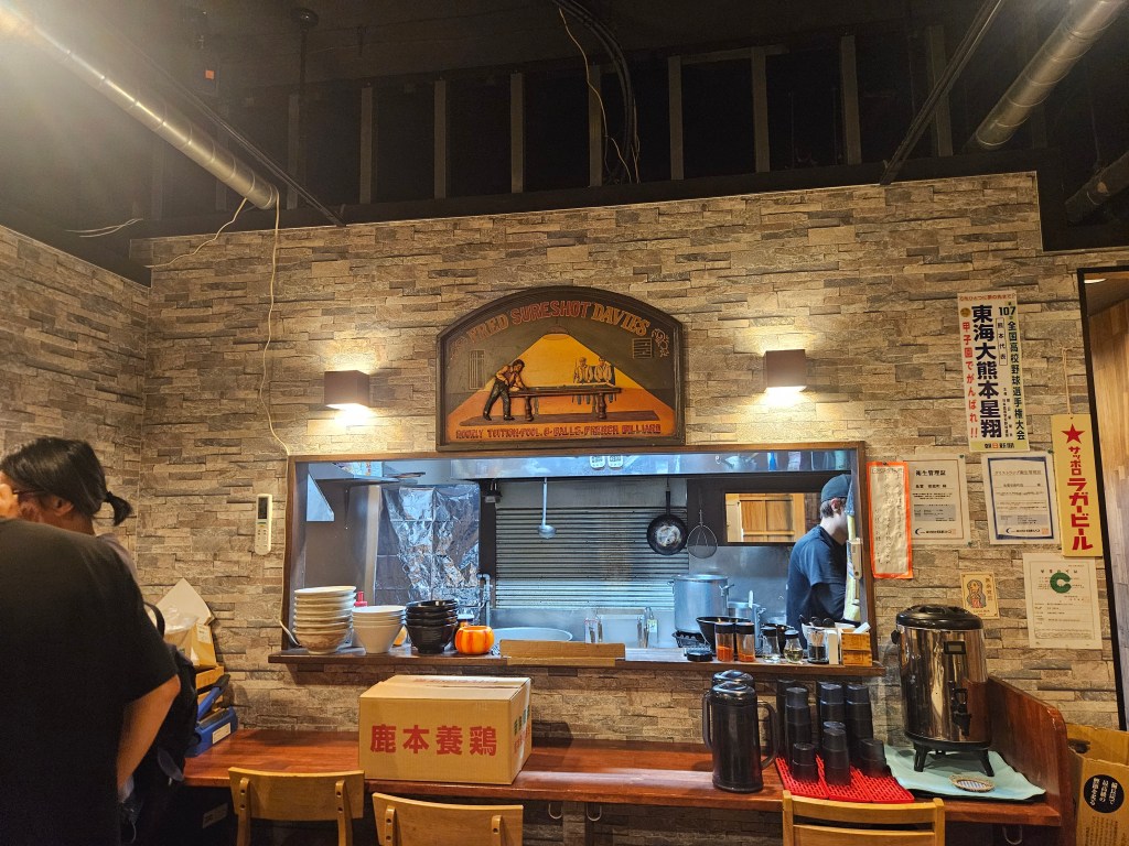 Intérieur d'un restaurant avec des murs en pierre, affiches et comptoir où un chef prépare des plats. Des assiettes et tasses sont visibles sur la table.