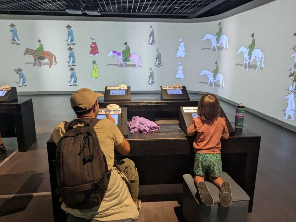 Une personne assise à un bureau dans un musée interactif, utilisant un écran tactile, tandis qu'un enfant se tient debout à un autre bureau, regardant des animations projetées sur les murs.