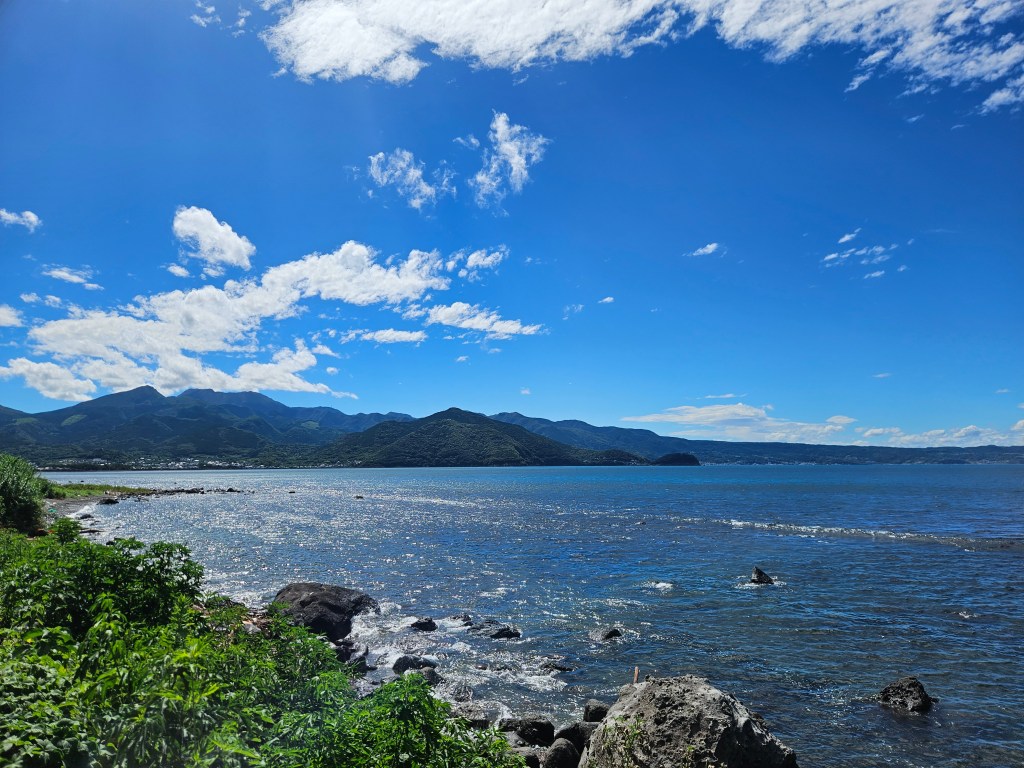 Vue panoramique sur la mer avec des montagnes en arrière-plan sous un ciel bleu parsemé de nuages blancs.