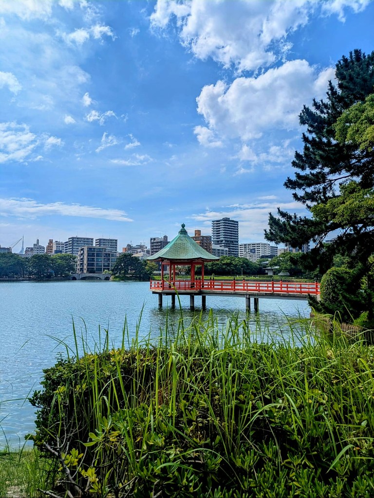 Vue d'un pavillon traditionnel sur pilotis au bord d'un lac, avec un ciel bleu parsemé de nuages et des immeubles en arrière-plan.