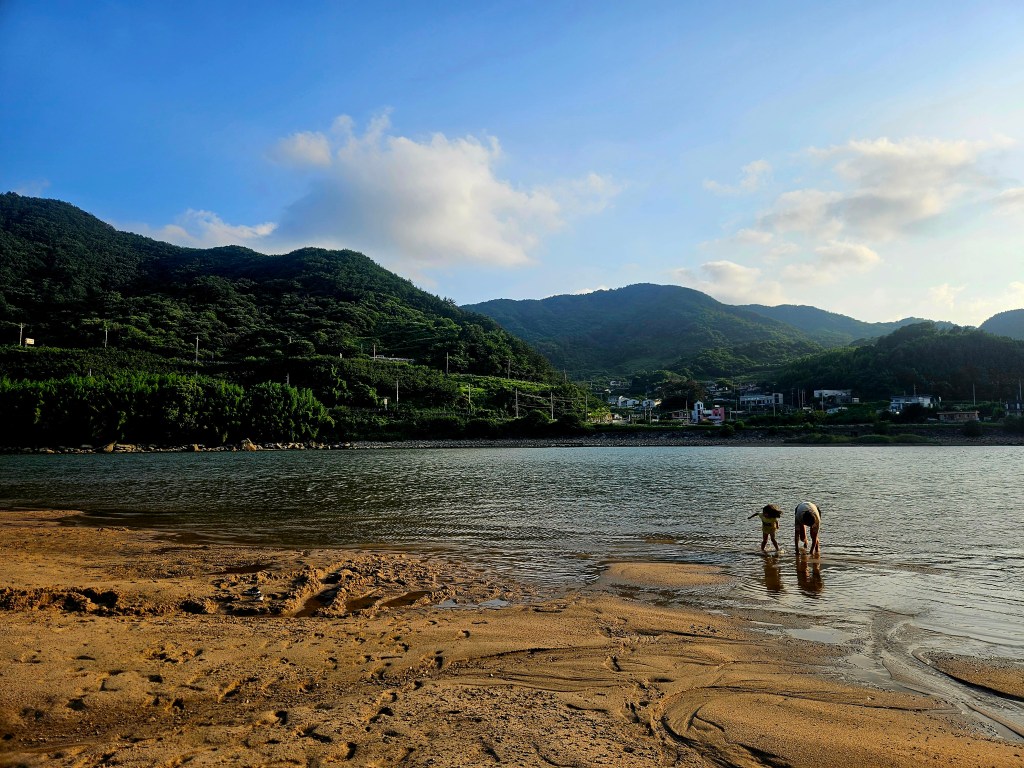 Deux personnes, un adulte et un enfant, jouent au bord d'un fleuve sous un ciel bleu avec des nuages, entourés de collines verdoyantes.