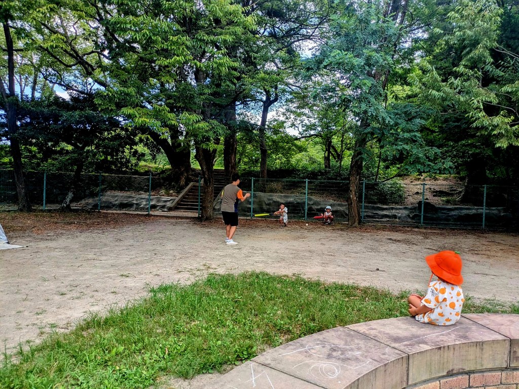 Des enfants jouent dans un parc. Un enfant en t-shirt gris et un short joue au baseball, tandis qu'un autre enfant est assis sur une petite marche, portant un chapeau orange. En arrière-plan, on aperçoit des arbres verdoyants et une clôture.