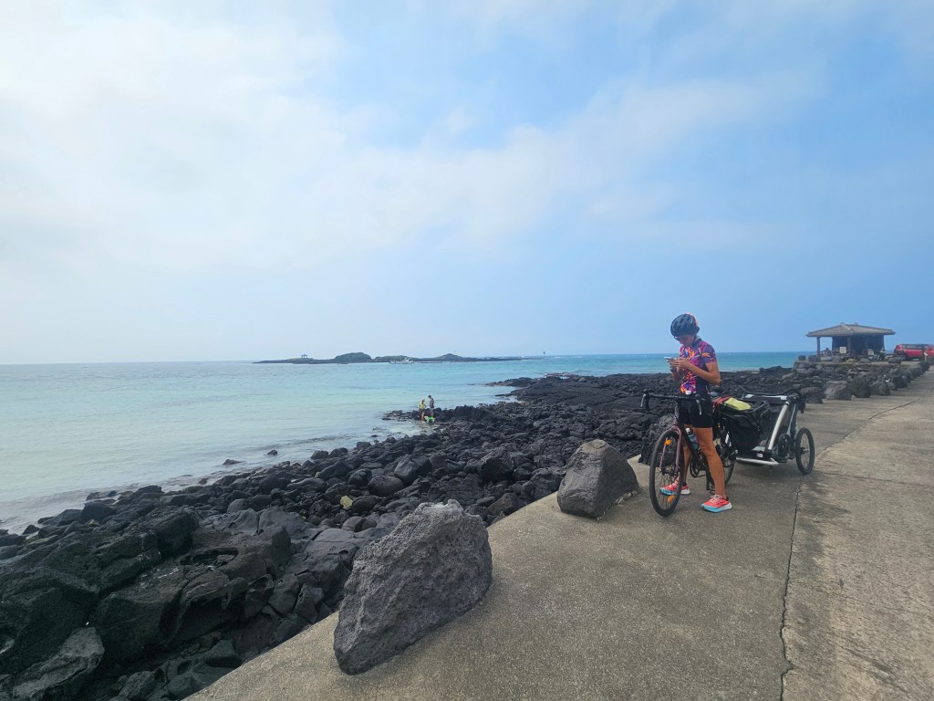 Femme en tenue de vélo regardant son téléphone sur un chemin en bord de mer, avec des rochers noirs et une eau turquoise en arrière-plan.