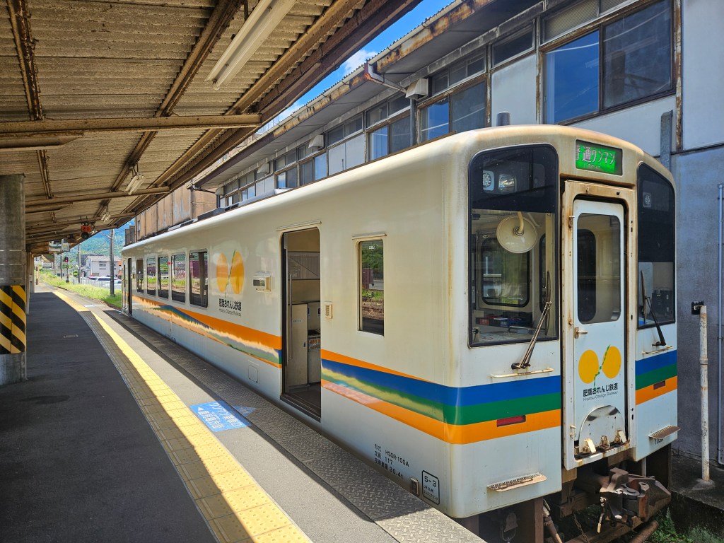 Un train de style japonais garé à une station, montrant sa carrosserie blanche avec des bandes colorées oranges, vertes et bleues, sous un ciel clair.