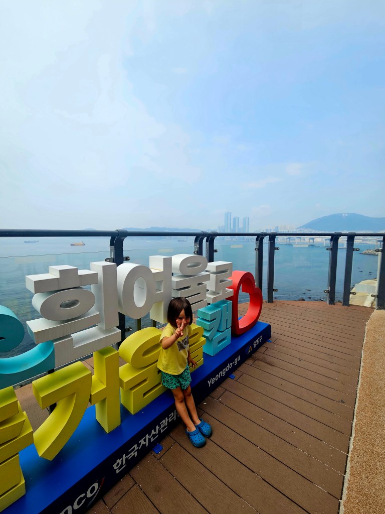 Une petite fille faisant un signe de paix devant un panneau coloré à Yeongdo, avec une vue sur la baie et des bâtiments en arrière-plan.